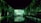 A dark, immersive space with vivid green projections covering the walls and ceiling, resembling a natural, forest-like setting. Silhouettes of people stand on a bridge, creating reflections in the water below.