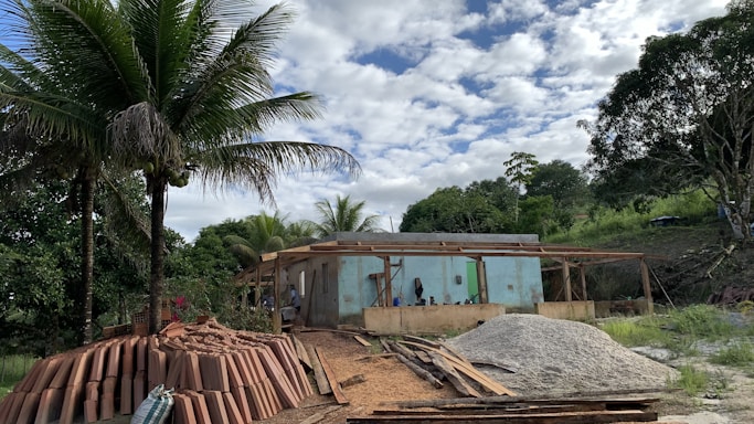 A construction site is set in a lush, green area with tall palm trees, indicating a tropical location. In the foreground, there are stacks of clay bricks and piles of sand and gravel. A partially built concrete structure with wooden scaffolding is visible, surrounded by dense vegetation and under a partly cloudy sky.