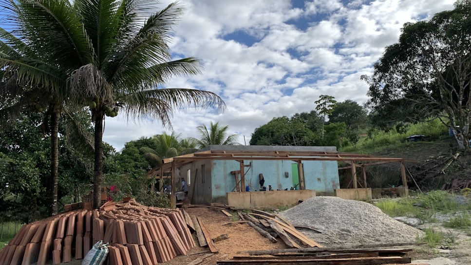 A construction site is set in a lush, green area with tall palm trees, indicating a tropical location. In the foreground, there are stacks of clay bricks and piles of sand and gravel. A partially built concrete structure with wooden scaffolding is visible, surrounded by dense vegetation and under a partly cloudy sky.