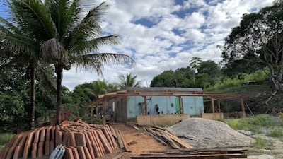 A construction site is set in a lush, green area with tall palm trees, indicating a tropical location. In the foreground, there are stacks of clay bricks and piles of sand and gravel. A partially built concrete structure with wooden scaffolding is visible, surrounded by dense vegetation and under a partly cloudy sky.