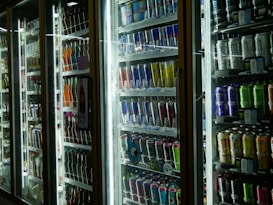 Brightly lit refrigerators filled with an assortment of beverages, including energy drinks, sodas, and juices, neatly arranged and displayed through glass doors.