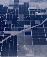 An aerial view of vast agricultural fields organized in a grid-like pattern. The fields are divided by straight roads and irrigation channels, creating rectangular sections. The landscape appears flat, with variations in color indicating different crops or land usage.