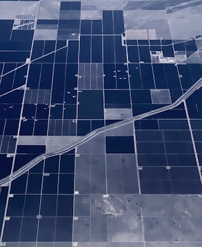 An aerial view of vast agricultural fields organized in a grid-like pattern. The fields are divided by straight roads and irrigation channels, creating rectangular sections. The landscape appears flat, with variations in color indicating different crops or land usage.