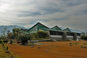 A modern building with a green roof and large glass windows, set against a backdrop of hazy mountains. The surrounding area is landscaped with various trees and shrubs, and the ground is covered with brownish grass. The sky is overcast with a hint of sunlight breaking through.
