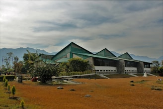 A modern building with a green roof and large glass windows, set against a backdrop of hazy mountains. The surrounding area is landscaped with various trees and shrubs, and the ground is covered with brownish grass. The sky is overcast with a hint of sunlight breaking through.