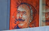 A mural depicting a man with glasses and a mustache, painted on a bright orange background. Part of the mural is reflected in a glass window, adding depth to the image. The intricate brushwork highlights facial features, giving the artwork a three-dimensional effect.