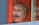 A mural depicting a man with glasses and a mustache, painted on a bright orange background. Part of the mural is reflected in a glass window, adding depth to the image. The intricate brushwork highlights facial features, giving the artwork a three-dimensional effect.