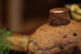 A loaf of bread is topped with a small, ornate wooden cup that appears to contain a creamy white substance. The surface of the bread is textured and studded with grains. In the background, there are hints of a green plant and lemon wedges, giving a cozy and rustic feel.