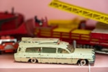 A close-up of a vintage toy ambulance car, slightly worn with chipped paint, resting on a pink surface. The toy is primarily cream-colored with some red detailing, and a red beacon light on the top. In the background, there is a blurred red and yellow structure, possibly another toy.