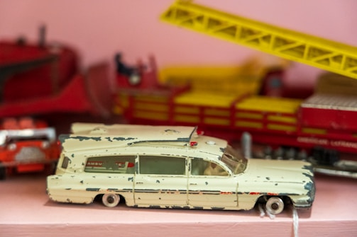A close-up of a vintage toy ambulance car, slightly worn with chipped paint, resting on a pink surface. The toy is primarily cream-colored with some red detailing, and a red beacon light on the top. In the background, there is a blurred red and yellow structure, possibly another toy.