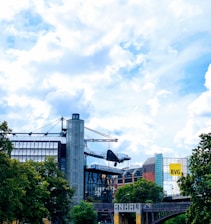 A commercial or historical aircraft is mounted as a display on top of a building in an urban area. Surrounding the site are modern architecture, trees, and a partly cloudy sky. The building features glass and metal design elements, and there is a yellow sign with the letters 'BVG' visible in the background.