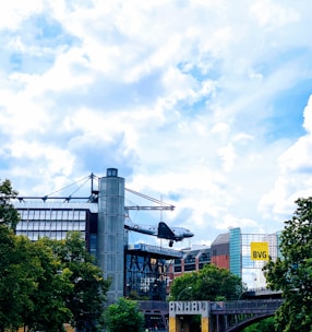 A commercial or historical aircraft is mounted as a display on top of a building in an urban area. Surrounding the site are modern architecture, trees, and a partly cloudy sky. The building features glass and metal design elements, and there is a yellow sign with the letters 'BVG' visible in the background.