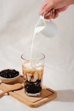 A hand is pouring milk from a small white pitcher into a tall glass filled with a beverage that has black tapioca pearls at the bottom and caramel-colored streaks mixing with the milk. Beside the glass, there is a wooden tray with a bowl of tapioca pearls and a wooden spoon.