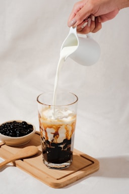 A hand is pouring milk from a small white pitcher into a tall glass filled with a beverage that has black tapioca pearls at the bottom and caramel-colored streaks mixing with the milk. Beside the glass, there is a wooden tray with a bowl of tapioca pearls and a wooden spoon.