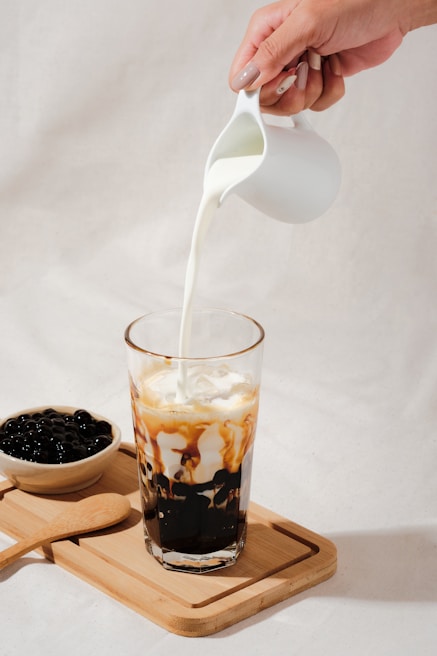 A hand is pouring milk from a small white pitcher into a tall glass filled with a beverage that has black tapioca pearls at the bottom and caramel-colored streaks mixing with the milk. Beside the glass, there is a wooden tray with a bowl of tapioca pearls and a wooden spoon.