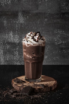 A tall glass filled with a rich chocolate milkshake topped with whipped cream and chocolate syrup. The drink sits on a round wooden board with scattered chocolate pieces surrounding it, set against a textured dark background.