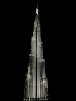 A tall, illuminated skyscraper with a sleek, modern design stands against a dark, black background. The building is architecturally impressive, featuring multiple tiers and a spire at the top, all highlighted with bright lighting.