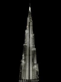 A tall, illuminated skyscraper with a sleek, modern design stands against a dark, black background. The building is architecturally impressive, featuring multiple tiers and a spire at the top, all highlighted with bright lighting.