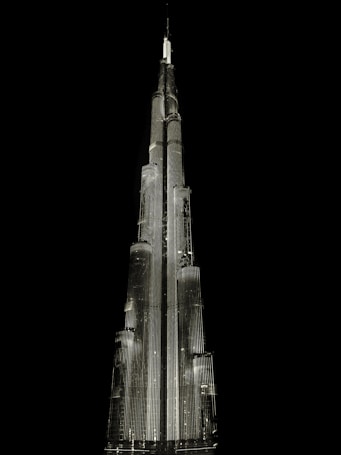 A tall, illuminated skyscraper with a sleek, modern design stands against a dark, black background. The building is architecturally impressive, featuring multiple tiers and a spire at the top, all highlighted with bright lighting.