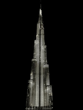 A tall, illuminated skyscraper with a sleek, modern design stands against a dark, black background. The building is architecturally impressive, featuring multiple tiers and a spire at the top, all highlighted with bright lighting.