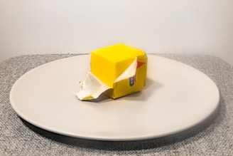 A cube of butter partially unwrapped from a yellow paper wrapping, placed on a large round white plate resting on a gray textured surface.