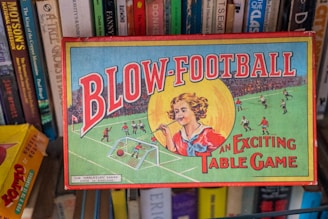 A vintage board game box titled 'Blow-Football' featuring colorful illustrations. The design includes a young person blowing a ball towards a miniature football goal, surrounded by players on a field. Shelves with various books are visible behind the box.