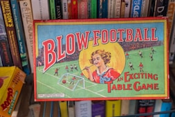 A vintage board game box titled 'Blow-Football' featuring colorful illustrations. The design includes a young person blowing a ball towards a miniature football goal, surrounded by players on a field. Shelves with various books are visible behind the box.