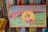 A vintage board game box titled 'Blow-Football' featuring colorful illustrations. The design includes a young person blowing a ball towards a miniature football goal, surrounded by players on a field. Shelves with various books are visible behind the box.