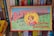 A vintage board game box titled 'Blow-Football' featuring colorful illustrations. The design includes a young person blowing a ball towards a miniature football goal, surrounded by players on a field. Shelves with various books are visible behind the box.