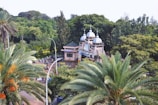Aerial view of a lush, green area featuring a building with white domes and several palm trees. The building appears to be of a religious nature, surrounded by dense foliage and people walking around its premises. A tall street lamp is visible, and the surroundings convey a serene, peaceful environment.
