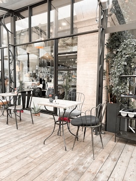 The image depicts an outdoor caf&eacute; with metal chairs and tables placed on a wooden deck. The area is enclosed by large glass windows, with a view of additional seating inside. Potted plants and ornamental lamps add to the ambiance, creating a cozy and inviting atmosphere.