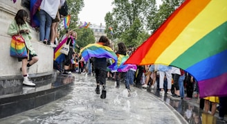 A lively outdoor event with many people holding and wearing rainbow flags, some individuals are running through a shallow pool of water, and others are standing by watching. The scene is bright and full of motion, with people looking joyful and engaged.