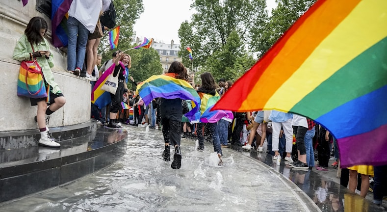 A lively outdoor event with many people holding and wearing rainbow flags, some individuals are running through a shallow pool of water, and others are standing by watching. The scene is bright and full of motion, with people looking joyful and engaged.