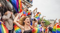 A vibrant crowd of people celebrating with rainbow flags, indicating a pride or LGBTQ+ event. The participants display various expressions of joy and excitement, some shouting and others taking pictures. The atmosphere seems lively and colorful.