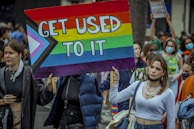 A group of people are gathered, with some holding a large sign featuring a colorful design that includes a rainbow and trans pride flag colors, saying 'Get Used To It'. The setting appears to be an outdoor march or demonstration, and some participants are wearing face masks.