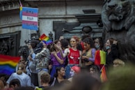 A group of people gather closely together in front of a large statue of a lion, holding rainbow flags and a sign with a pink, white, and blue flag pattern that reads 'PMA POUR TOU.TES'. Many in the crowd appear cheerful and are dressed in colorful attire. Some individuals are wearing masks, and the atmosphere is lively, indicating participation in a public event.