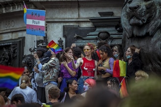 A group of people gather closely together in front of a large statue of a lion, holding rainbow flags and a sign with a pink, white, and blue flag pattern that reads 'PMA POUR TOU.TES'. Many in the crowd appear cheerful and are dressed in colorful attire. Some individuals are wearing masks, and the atmosphere is lively, indicating participation in a public event.