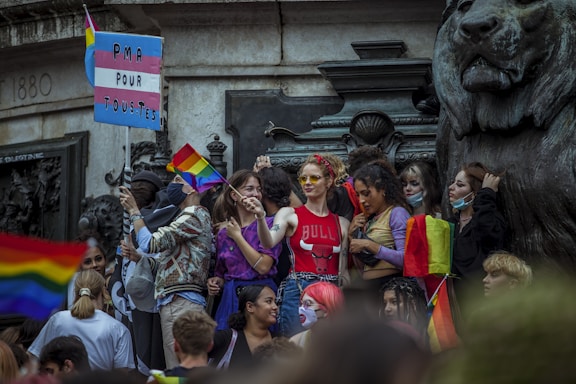 A group of people gather closely together in front of a large statue of a lion, holding rainbow flags and a sign with a pink, white, and blue flag pattern that reads 'PMA POUR TOU.TES'. Many in the crowd appear cheerful and are dressed in colorful attire. Some individuals are wearing masks, and the atmosphere is lively, indicating participation in a public event.
