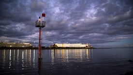 A large cargo ship with the words 'HOEGH AUTOLINERS' is docked near a harbor. The water reflects the lights from the ship and nearby structures. Dark, dramatic clouds fill the sky, and a navigational marker with a red sign and light is positioned in the foreground on the water.