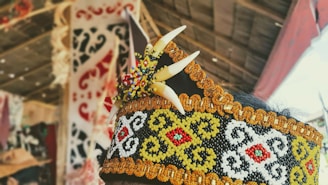 An ornate headpiece featuring intricate beadwork in black, white, yellow, and red patterns. It includes decorative elements resembling teeth and colorful beads. The background shows a blurred view of traditional crafts and fabrics.