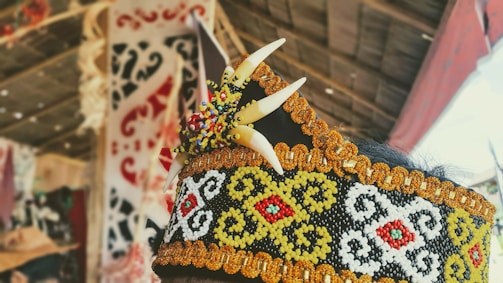 An ornate headpiece featuring intricate beadwork in black, white, yellow, and red patterns. It includes decorative elements resembling teeth and colorful beads. The background shows a blurred view of traditional crafts and fabrics.