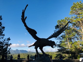 A large eagle sculpture with outstretched wings stands prominently against a backdrop of distant mountains and a clear blue sky. Several people are gathered near the statue, taking in the scenery. The surrounding area is lush with green trees, creating a natural setting.