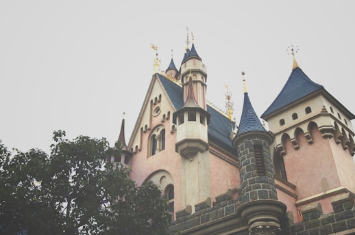 A fairytale-like castle with blue pointed roofs and intricate architectural details, featuring gold accents and surrounded by lush green foliage.