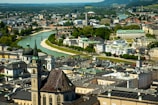 Aerial view of a vibrant cityscape featuring historic architecture with ornate spires and domes. The city is nestled along a river with bridges, surrounded by greenery and distant hills. The blend of old and modern buildings creates a dynamic and picturesque urban scene.