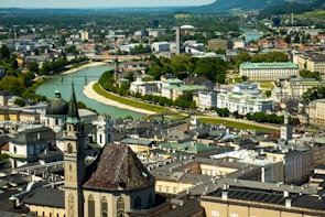 Aerial view of a vibrant cityscape featuring historic architecture with ornate spires and domes. The city is nestled along a river with bridges, surrounded by greenery and distant hills. The blend of old and modern buildings creates a dynamic and picturesque urban scene.