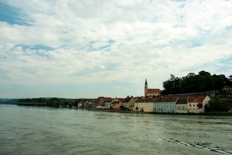 A picturesque riverside town with colorful historic buildings lining the waterfront. A church tower with a clock is prominently visible among the structures. The sky is partly cloudy, and lush greenery is visible in the background.