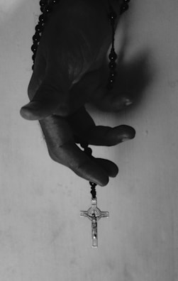 A hand gently holds a rosary, with a cross at the end of the beads. The image is in black and white, emphasizing the texture of the hand and the smoothness of the beads.