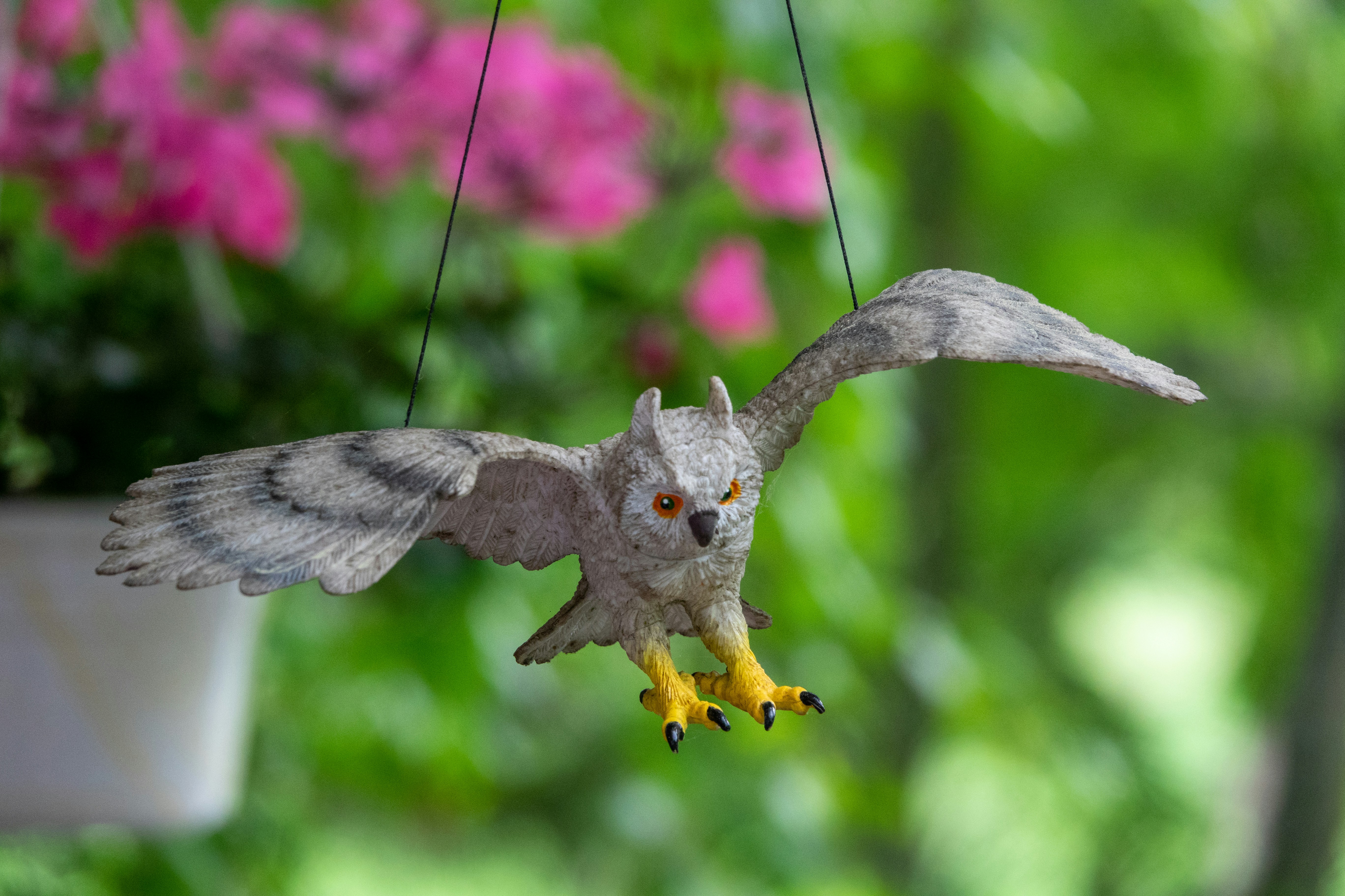 Gray owl model suspended by thin cords hovers before a lush, blurred garden backdrop.