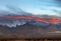 A breathtaking mountain range with snow-capped peaks under a dramatic sky. The sunset casts vibrant orange and red hues across the clouds, contrasting against the cooler tones of the rugged mountains below.