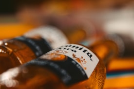 Close-up view of two brown glass bottles lying on their sides. The focus is on the labels, with details slightly blurred in the background. The foreground bottle's label displays the text clearly, offering a sense of intimacy and casual atmosphere.